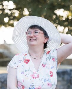 Portrait de Florence [Nom], traductrice spécialisée en théologie et en œcuménisme, portant une robe fleurie, une capeline blanche et des lunettes roses, souriant en extérieur sous des arbres.